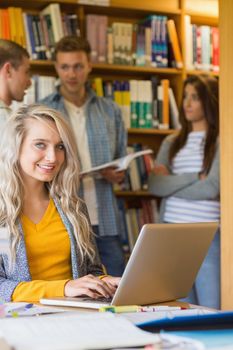 Portrait of a smiling female student using laptop while others in background at the college library