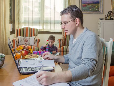 Father working hard in home office with notebook and his boring son playing on the background