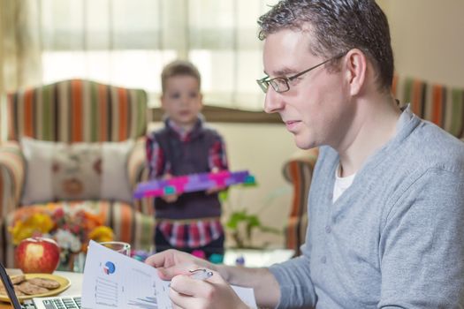 Father working hard in home office with notebook and his boring son playing on the background