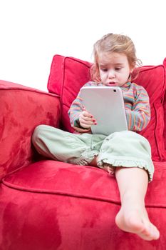Cute little girl sitting barefoot on a red sofa with a tablet computer surfing the internet with a quaint look of awe and concentration