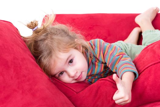 Beautiful little girl giving a thumbs up gesture as she lies comfortably on her stomach on a red couch looking at the camera with a friendly smile