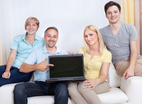 Smiling father surrounded by his happy teenage family pointing to a blank laptop screen for your advertising and marketing