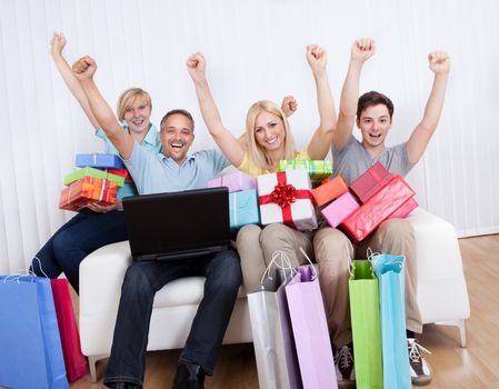 Happy family of online shoppers laden with gifts sitting around the father who is sitting with his laptop computer on his lap