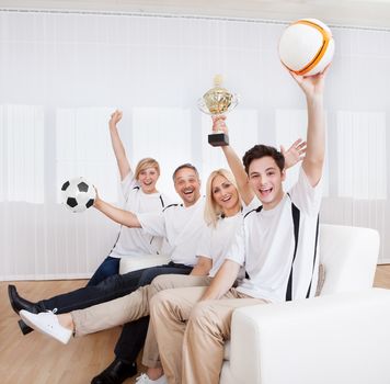 Ecstatic family sitting together on a couch celebrating a win with their arms raised in jubilation holding aloft a ball and trophy