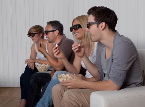 Family with teenage children sitting together on a couch eating bowls of popcorn wearing 3d glasses and watching the television
