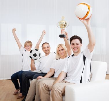 Ecstatic family sitting together on a couch celebrating a win with their arms raised in jubilation holding aloft a ball and trophy