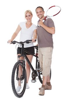Fit healthy middle-aged parents who enjoy an active outdoor lifestyle standing with a tennis racquet and bicycle