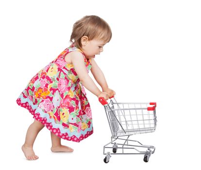 Cute little barefoot girl in a colourful dress pushing a miniature wire shopping trolley isolated on white