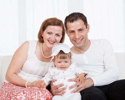 Proud young parents posing sitting on a sofa with their laughing little baby daughter