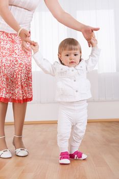 Cute little blonde baby girl taking her first steps in the nursery aided and supported by her mother