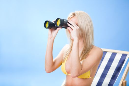 Beautiful young woman in bikini lying on a deckchair looking through binoculars at the beach