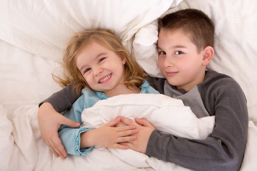 Mischievous young brother and sister in bed grinning up at the camera as they lie side by side under the duvet preparing to go to sleep