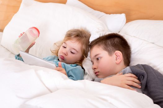 Cute little boy and girl reading a bedtime story on a tablet-pc before sleeping, lying in a big comfortable bed together