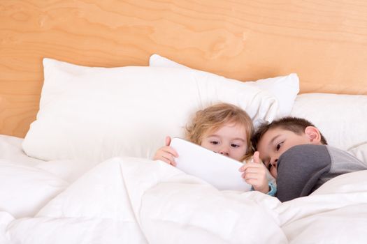 Little brother and sister snuggling up together at bedtime under a warm duvet as they read an e-book or surf the internet on a tablet-pc