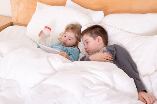 Happy little brother and sister lying in bed together as they cuddle under the duvet reading a tablet computer before going to sleep