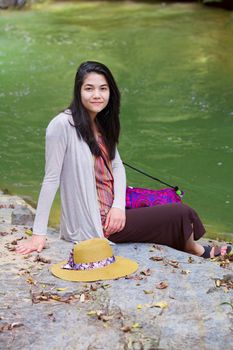 Biracial teen girl sitting next to river, smiling