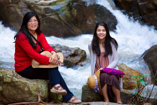 Asian mother and daughter sitting on boulder by riverside
