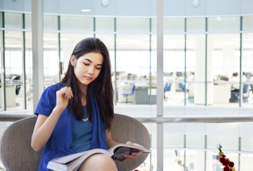 Beautiful biracial teen girl reading magazine in office