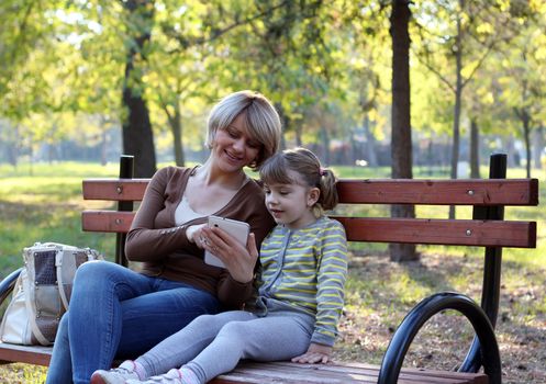 mother and daughter sitting on bench and play with tablet