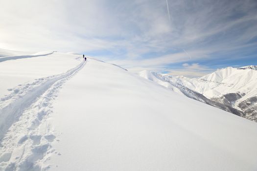 Alpinist hiking uphill by ski touring on the mountain ridge in powder snow with deep track in the foreground and scenic high mountain view in the background