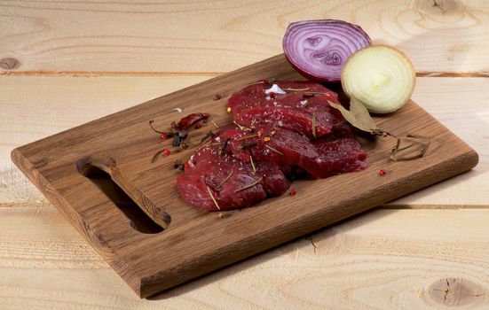 Arrangement of Raw Sliced Beef with Halves of White and Red Onion, Bay Leafs and Spices on Cutting Board isolated on Wooden background