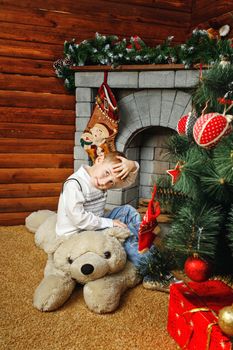 Boy sitting on teddy bear next to Christmas tree and gifts on background of fireplaces