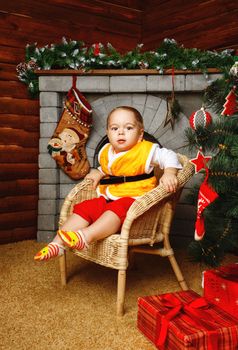 Little boy sitting in wicker chair near Christmas tree on background of fireplaces