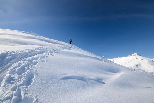 Alpinist hiking uphill by ski touring on the mountain ridge in powder snow with deep track in the foreground and scenic high mountain view in the background