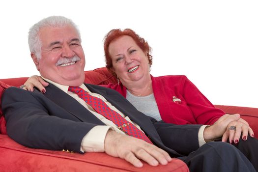 Caucasian senior couple smiling and holding hands, wearing elegant clothes, while sitting on a comfortable red couch