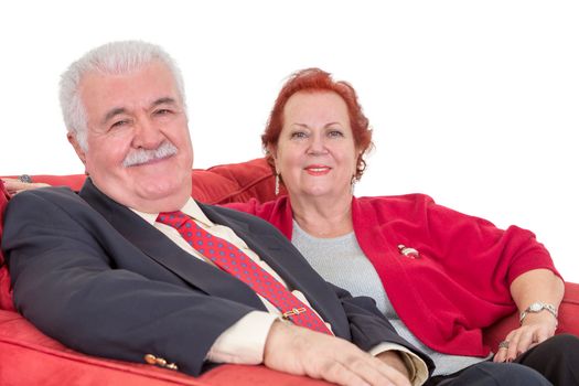 Stylish senior couple in colour co-ordinated red clothing sitting together on a red sofa smiling at the camera, close up view over white