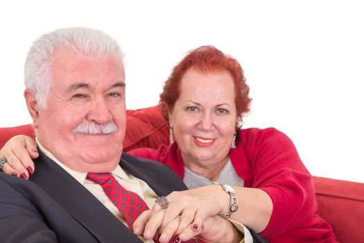 Affectionate senior couple sitting close together holding hands on a red sofa and looking at the camera with contented friendly smiles, close up view on white