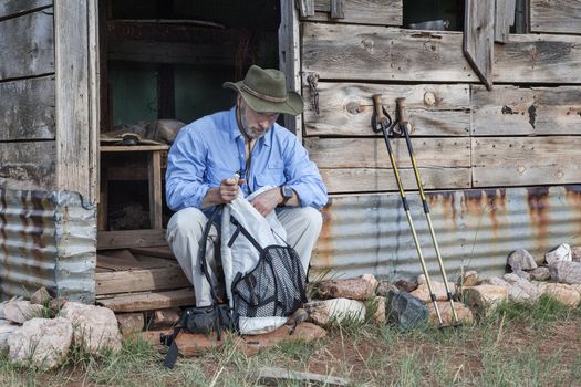 senior male hiker sitting with a backpack and trekking poles in front of old mountain cabin or shack in Colorado