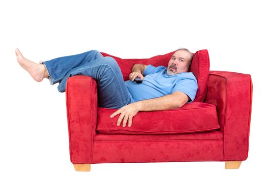 Man engrossed in watching television lying barefoot on a red couch with the remote control in his hand and a look of fascinated concentration, isolated on white
