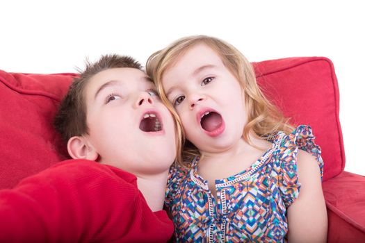 Two playful young children pulling faces as an attractive brother and sister sit together on a red couch playing copycat with their mouths wide open