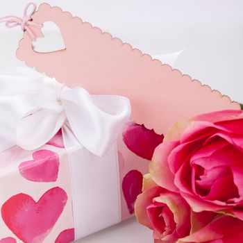 Close-up of a gift box wrapped in decorative paper with pattern with heart shapes, with an empty tag, next to three roses, symbol of love and appreciation