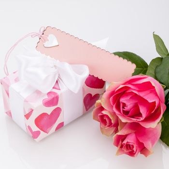 Close-up of a gift box wrapped in decorative paper with pattern with heart shapes, with an empty tag, next to three roses, symbol of love and appreciation