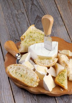 Arrangement of Delicious Camembert, Brie and Parmesan Cheese with Garlic and Herbs Bread and Cheese Knifes on Cheese Board closeup on Rustic Wood background