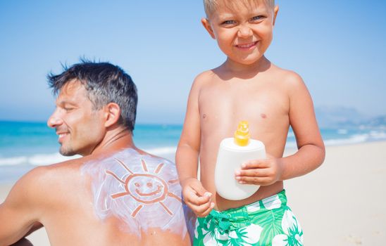 Adorable boy at tropical beach applying sunblock cream on a father's back.