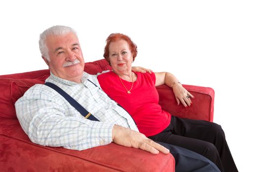 Attractive elderly couple smiling in satisfaction as they sit arm in arm on a comfortable red sofa over a white background