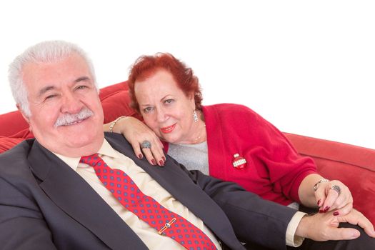 Stylish elderly couple sitting close together side by side on a red sofa looking sideways at the camera with a smile over a white background