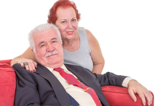 Loving elderly couple relaxing together on a red couch with the wife sitting behind her husband with her arm around his shoulders looking at the camera