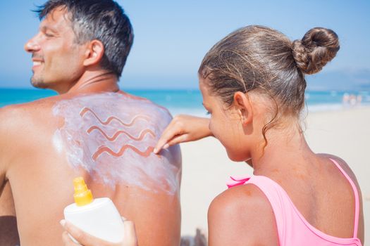 Adorable girl at tropical beach applying sunblock cream on a father's back.