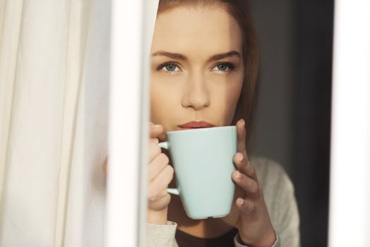 Beautiful caucasian woman drinking hot coffee or tea and looking through window. Indoor background.