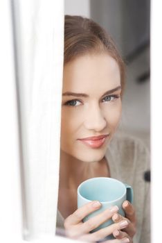 Young beautiful woman holding a cup and looking through window. Indoor background.