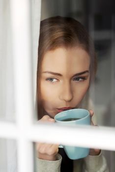 Beautiful caucasian woman drinking hot coffee or tea and looking through window. Indoor background.