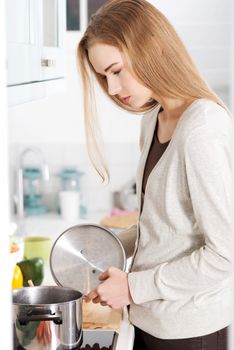 Beautiful caucasian woman is cooking standing in the kitchen and looking into kitchen pot.