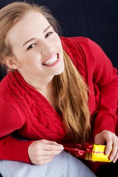 Beautiful caucasian woman in red sweater is unwrappind small present and sitting on a sofa.