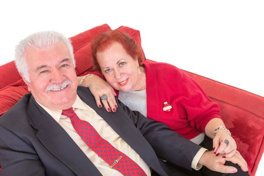 Senior couple sitting on a comfortable red couch holding hands affectionately and looking up at the camera with friendly smiles