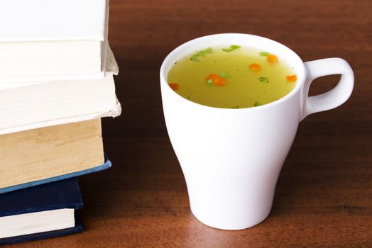 Hot vegetable soup in a cup on table next to books.