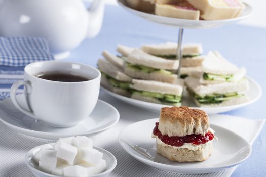 Cup of tea with sugar cubes and scones, cucumber sandwiches in the background